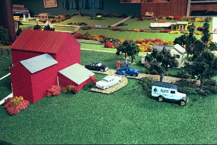 Big Red barn in foreground with farm house behind some trees, model depicting Glenn's home in the 1950s.